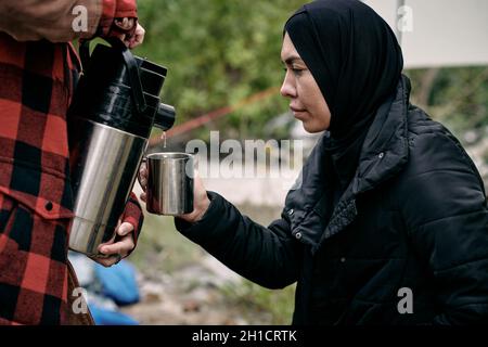 Junge muslimische Flüchtlingsfrau in schwarzen Kleidern mit metallischem Becher, während sie freiwillig heißes Wasser gießt Stockfoto