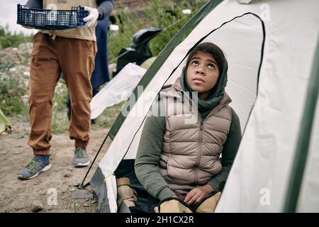 Afrikanisches Kind in Casualwear, das im Zelt sitzt und mit einer freiwilligen Tragetasche mit Futter auf dem Hintergrund nach oben blickt Stockfoto