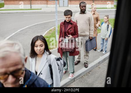 Verschiedene Gruppen von Menschen stehen in der Schlange an der Bushaltestelle mit sozialer Distanzierung und Eintritt in den Bus Stockfoto