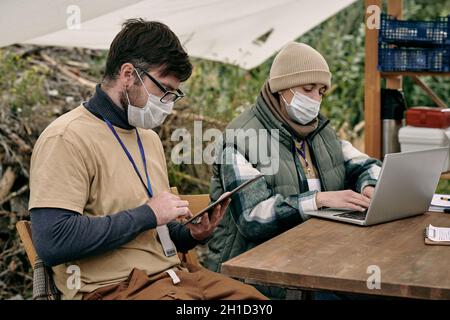 Zwei junge Freiwillige mit mobilen Geräten arbeiten mit Daten am Tisch im Spendenzelt Stockfoto