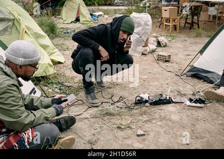 Zwei junge männliche Flüchtlinge, die Smartphones benutzen, während sie in ihrem Lager auf dem Boden sitzen Stockfoto