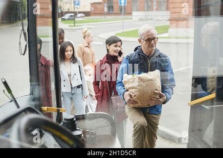 Durch den Blick aus Glas auf eine Gruppe von Menschen, die an der Bushaltestelle Schlange stehen und in den Bus einsteigen Stockfoto