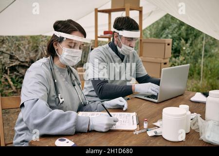 Zwei junge Freiwillige, die während ihrer Arbeit im Flüchtlingslager medizinische Dokumente ausfüllen und Daten in die Datenbank eingeben Stockfoto