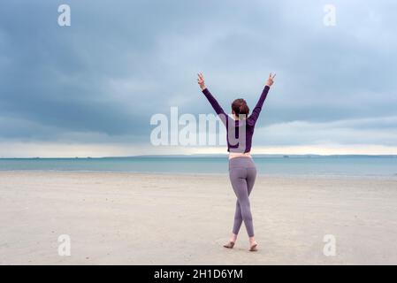 Junge Frau in Sportkleidung steht und hebt die Arme für die Freiheit am Strand Stockfoto