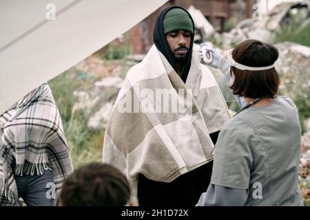 Ärztin mit elektronischem Thermometer zur Messung der Körpertemperatur des kranken Mannes im Flüchtlingslager Stockfoto