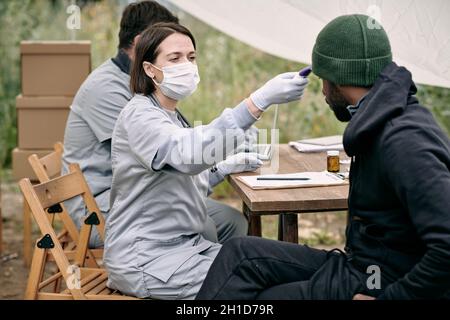 Arzt in Schutzmaske zur Messung der Körpertemperatur des kranken Mannes aus dem Migrationslager Stockfoto