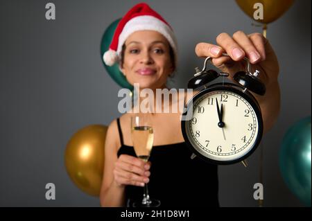 Konzentrieren Sie sich auf einen schwarzen Wecker mit Mitternacht auf dem Zifferblatt in der Hand einer fröhlichen Frau, die einen Weihnachtsmütze trägt und eine Sektflöte auf festlichem Grau hält Stockfoto