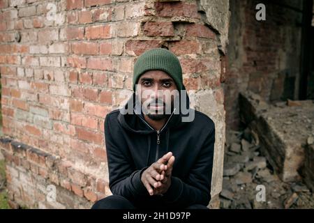 Junger afrikanischer Mann in Kapuzenpullover und Mütze, der an einer Ziegelwand eines zerstörten Gebäudes sitzt Stockfoto