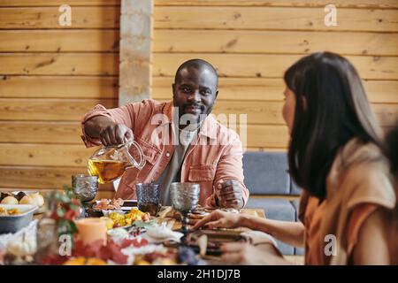 Afrikanischer junger Mann goss den Saft ins Glas und sprach mit der Frau am Esstisch Stockfoto
