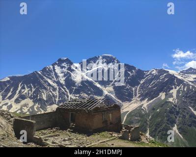 Altes verlassenes Haus in den Bergen. Ruinen einer Hütte ohne Dach auf dem Hintergrund der schneebedeckten Gipfel der Berge. Stockfoto