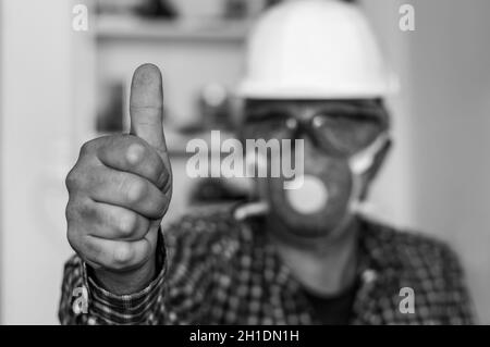 Arbeiter zeigt Daumen nach oben, mit Hut, Schutzbrille und Schutzmaske, selektiver Fokussierung. Stockfoto