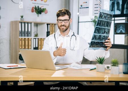 Kaukasischer Arzt lächelt mit dem Daumen nach oben auf der Kamera, während er am Schreibtisch sitzt und Tomographie-Scans in den Händen hält. Bärtiger Arzt mit Brille, weißem Laborkittel und Laptop im Büro. Stockfoto