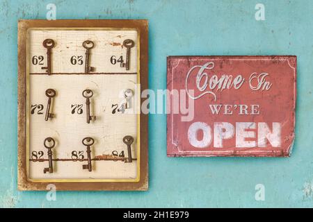 Kleines Vintage-Kabinett mit verrosteten Hotelschlüsseln und Zimmernummern und kommen herein wir sind offenes Schild auf einem blau erodierten Hintergrund Stockfoto