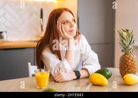 Seitenansicht von träumenden lächelnden attraktiven rothaarige junge Frau, die am Tisch mit exotischen tropischen Früchten in der Küche mit heller moderner Einrichtung sitzt, Stockfoto