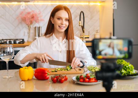 Mittellange Aufnahme Porträt der attraktiven Rotschopf Frau vlogger schießen Video Lebensmittel Blog über das Kochen auf Kamera des Mobiltelefons Stockfoto