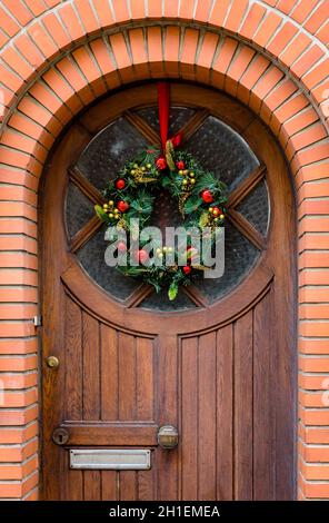 Vorderansicht einer Weihnachtskrone aus Kunststoff-Tannenzweigen mit Kugeln und Tannenzapfen, die an einer halb verglasten hölzernen Haustür mit einem gemauerten fram hängen Stockfoto