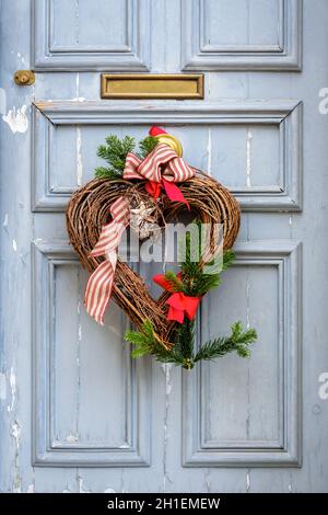 Vorderansicht einer herzförmigen Weihnachtsdekoration aus Ästen und Bändern, die an einer abgenutzten blauen Haustür mit Zierleisten hängt. Stockfoto