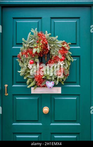 Vorderansicht einer Weihnachtskrone aus Tannenzweigen, Lametta und Kugeln, die an einer grünen Haustür mit Zierleisten hängt. Stockfoto