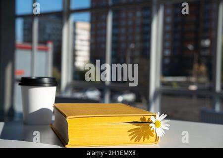 Geschlossenes gelbes Buch mit einem Lesezeichen mit einer Kamillenblüte und aromatischem Kaffee in einer Einweg-Papiertasse liegen auf dem Tisch in einem Café. Stockfoto