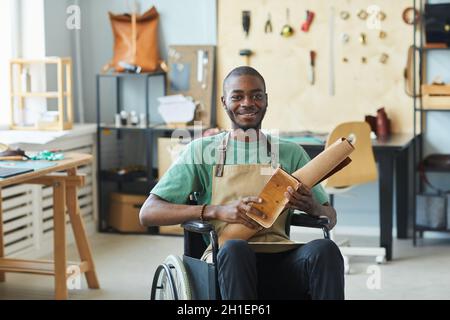 Porträt eines lächelnden afroamerikanischen Mannes, der im Rollstuhl sitzt und in der Lederwerkstatt arbeitet und Kamera und Kopierraum ansieht Stockfoto
