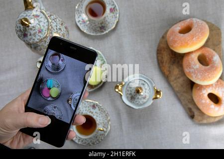 Draufsicht Bild der Dame, Blogger sitzen im Café und machen Foto mit Mobile von Lebensmitteln, Nachmittagstee mit Donuts und Makronen auf dem Tisch hoch Stockfoto