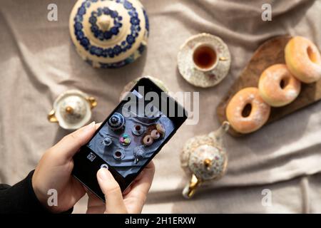 Draufsicht Bild der Dame, Blogger sitzen im Café und machen Foto mit Mobile von Lebensmitteln, Nachmittagstee mit Donuts und Makronen auf dem Tisch hoch Stockfoto