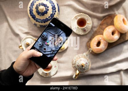 Draufsicht Bild der Dame, Blogger sitzen im Café und machen Foto mit Mobile von Lebensmitteln, Nachmittagstee mit Donuts und Makronen auf dem Tisch hoch Stockfoto