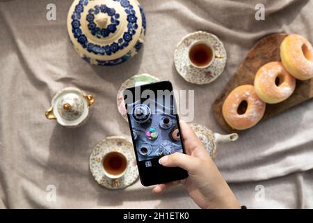 Draufsicht Bild der Dame, Blogger sitzen im Café und machen Foto mit Mobile von Lebensmitteln, Nachmittagstee mit Donuts und Makronen auf dem Tisch hoch Stockfoto