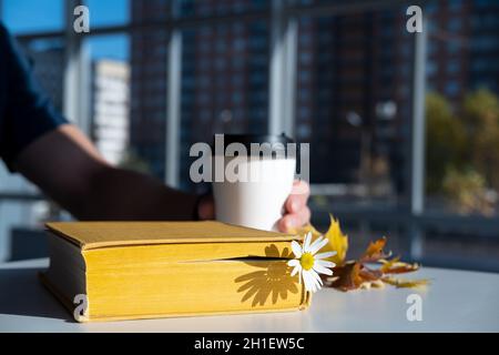 Buch mit einem Lesezeichen mit einer Kamillenblume liegt auf dem Tisch, vor dem Hintergrund hält eine männliche Hand eine Einweg-Tasse Kaffee. Stockfoto