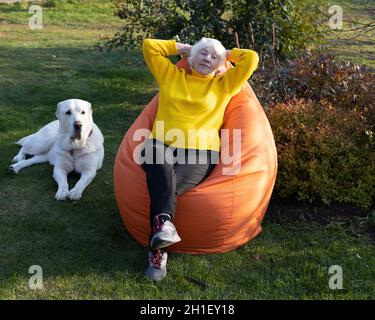 Die attraktive, grauhaarige 85-jährige Frau sitzt auf einem Taschenstuhl und hört Musik über Kopfhörer. Stockfoto