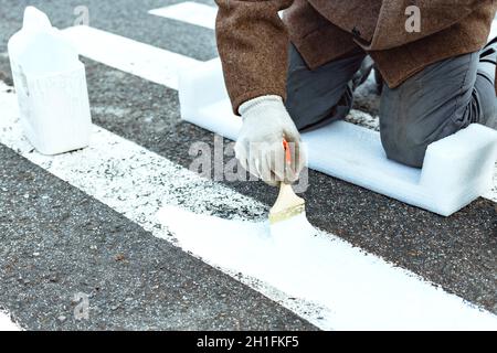 Ein Mann malt mit einem Pinsel auf die Asphaltmarkierungen für eine Fußgängerüberfahrt. Straßenreparatur. Stockfoto