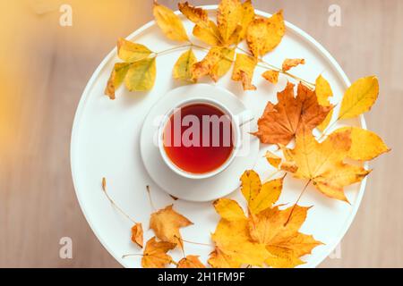 Duftende schwarze Teetasse auf weißem Couchtisch, gefallene gelbe Ahornblätter Stockfoto