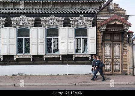 Zwei Frauen, die in Ulan-Ude an einem traditionellen Herrenhaus aus Holz mit geschnitzten Fenstern und Fensterläden vorbeigehen. Republik Burjatien, Russland Stockfoto