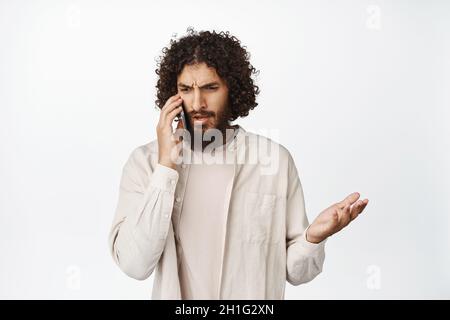 Frustrierter junger Mann aus dem Nahen Osten, der mit einem verworrenen Gesicht auf dem Mobiltelefon spricht, mit einem zuckenden Rüpselchen aufgeregt und auf weißem Hintergrund steht Stockfoto