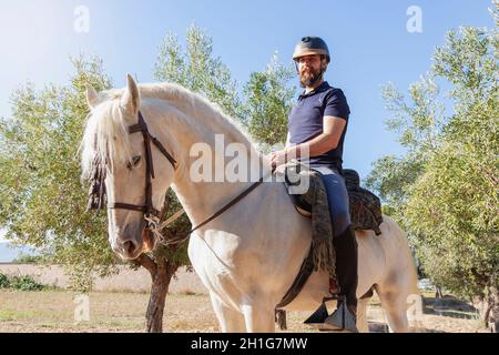 Ein behelmter Reiter, ein bärtiger Kaukasusmann, reitet auf seinem weißen Pferd auf einem Feld, umgeben von Olivenbäumen und blauem Himmel im Hintergrund. Stockfoto