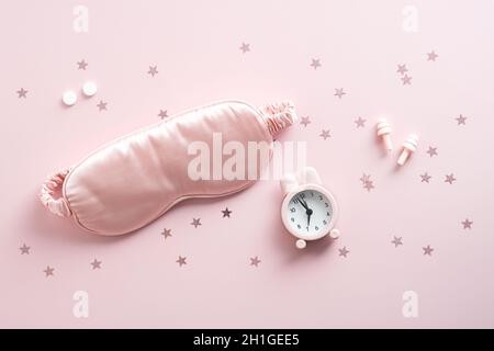 Weibliche schlafende Augenmaske, Wecker, Ohrstöpsel und Pillen auf rosa Hintergrund. Konzept zur Behandlung von Schlaflosigkeit. Stockfoto