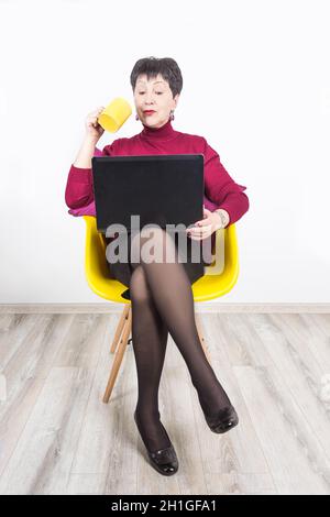 Ältere Frau mit Laptop und Tasse Kaffee auf grauem Studiohintergrund. Stockfoto