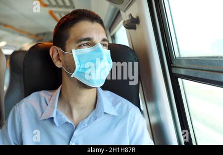 Fahren Sie sicher mit öffentlichen Verkehrsmitteln. Junger Mann mit OP-Maske, der durch das Zugfenster schaut. Zug Passagier mit Schutzmaske durch die Stockfoto