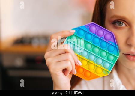 Nahaufnahme einer jungen Rothaarigen Frau, die in der Hand neben dem Gesicht farbenfrohe, schillernde, weiche Silikonblasen zu Hause hält. Stockfoto