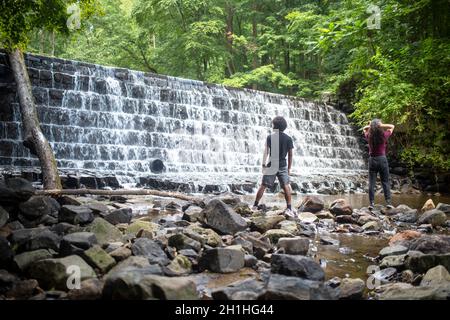 Wasserfällen über dem abgestuften Wasserfall mit üppigem Waldbelaub dahinter. Zwei junge Männer, afroamerikanisch und kaukasisch, stehen auf Steinen an der Basis Stockfoto