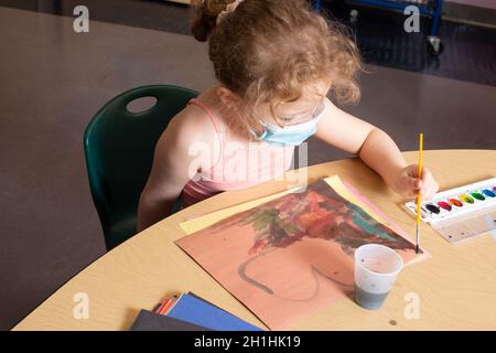 Bildung Vorschule 4-5-jähriges Mädchen, das am Tisch mit Wasserfarben sitzt, mit der linken Hand, Gesichtsmaske trägt Stockfoto