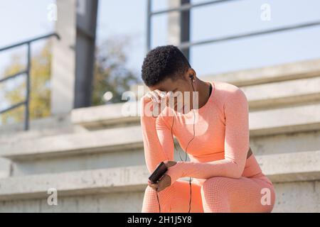 Seriöse junge Sportlerin in einem pinken Anzug, die sich nach dem Training ausruht, Musik und Podcast auf einem Mobiltelefon hört Stockfoto