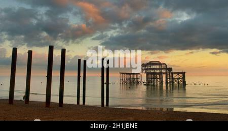 Der verwelkte West Pier bei einem bewölkten Sonnenuntergang, Brighton, England. Der Pier wurde 1866 erbaut und 1975 geschlossen. Er ist nach wie vor unter Denkmalschutz der Klasse I und ein bekanntes Wahrzeichen. Vögel strömen zu der rostenden Struktur. Säulen, die auf die strukturellen Überreste des alten Piers auf dem Pier hinweisen. Stockfoto