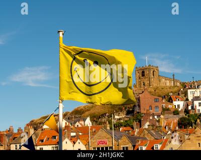 Gelbe Smiley-Flagge flattert im Wind an einem sonnigen Tag mit der St. Mary's Church, Whitby in der Ferne. North Yorkshire, Großbritannien Stockfoto