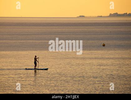 Ein silhouettierter Paddlebarder, der bei Sonnenuntergang an einem Hove Strand paddeln kann. Brighton & Hove, Engand, Großbritannien. Worthing Pier, West Sussex am Horizont. Stockfoto