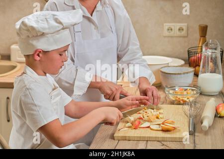 Mutter und Sohn in weißen Chefkleidern schnitten in ihrer eigenen Küche mit einem Messer Birnen. Eine Erwachsene Frau mit einem kleinen Jungen wird einen Kuchen kochen Stockfoto