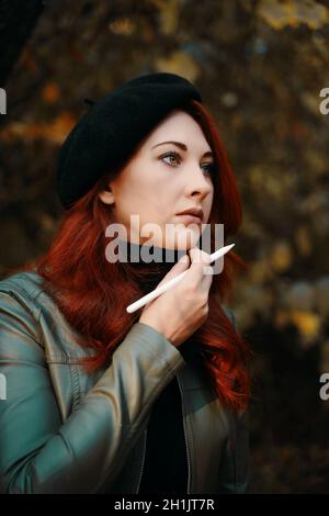 Schöne rothaarige Frau im Park. Brütende Frau in einer Baskenmütze und Lederjacke. Mann mit Stift, Designer, Freiberufler. Herbstporträt. Warten auf Inspiration. Stockfoto