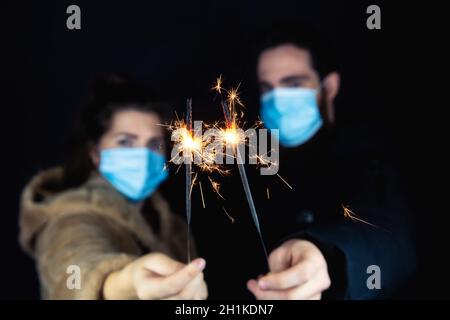 Junges Paar mit schützenden Gesichtsmasken für Coronavirus und mit Wunderkerzen für Silvester, 2021 und Covid-19 Konzept Stockfoto