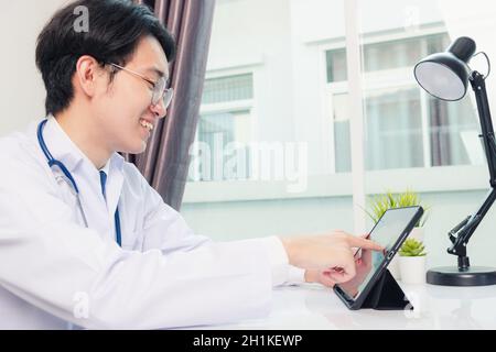 Glücklich asiatische junge Arzt Handsome Mann Lächeln Arbeit aus dem Home Office tragen Brille, mit schwarzen modernen smart digital Tablet-Computer auf dem Schreibtisch im Krankenhaus o Stockfoto