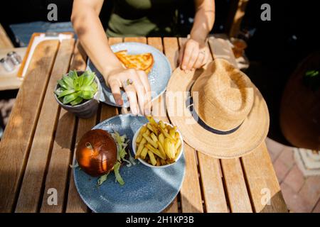 Bild von einem Tisch und den Händen einer Frau, die auf einem Holztisch nach einem Hamburger und Pommes streut Stockfoto